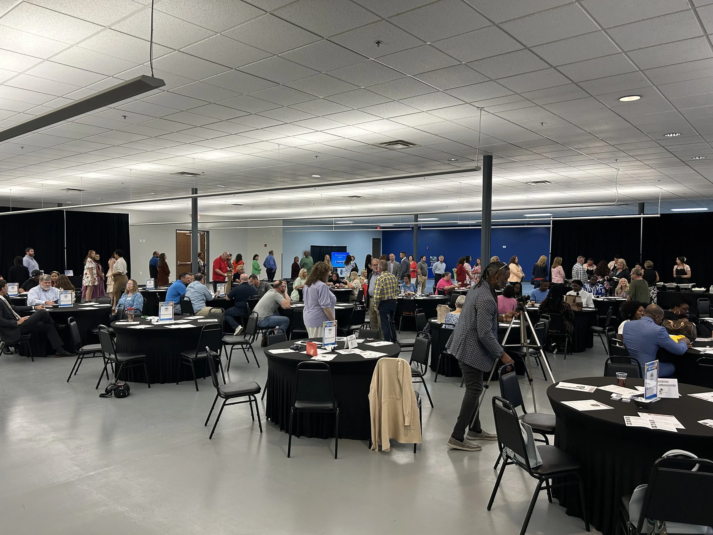 A large conference or event room filled with many tables and chairs, with people standing and sitting, engaged in conversations, and some working on laptops. The room has high ceilings with fluorescent lighting withblack pipe and drape.
