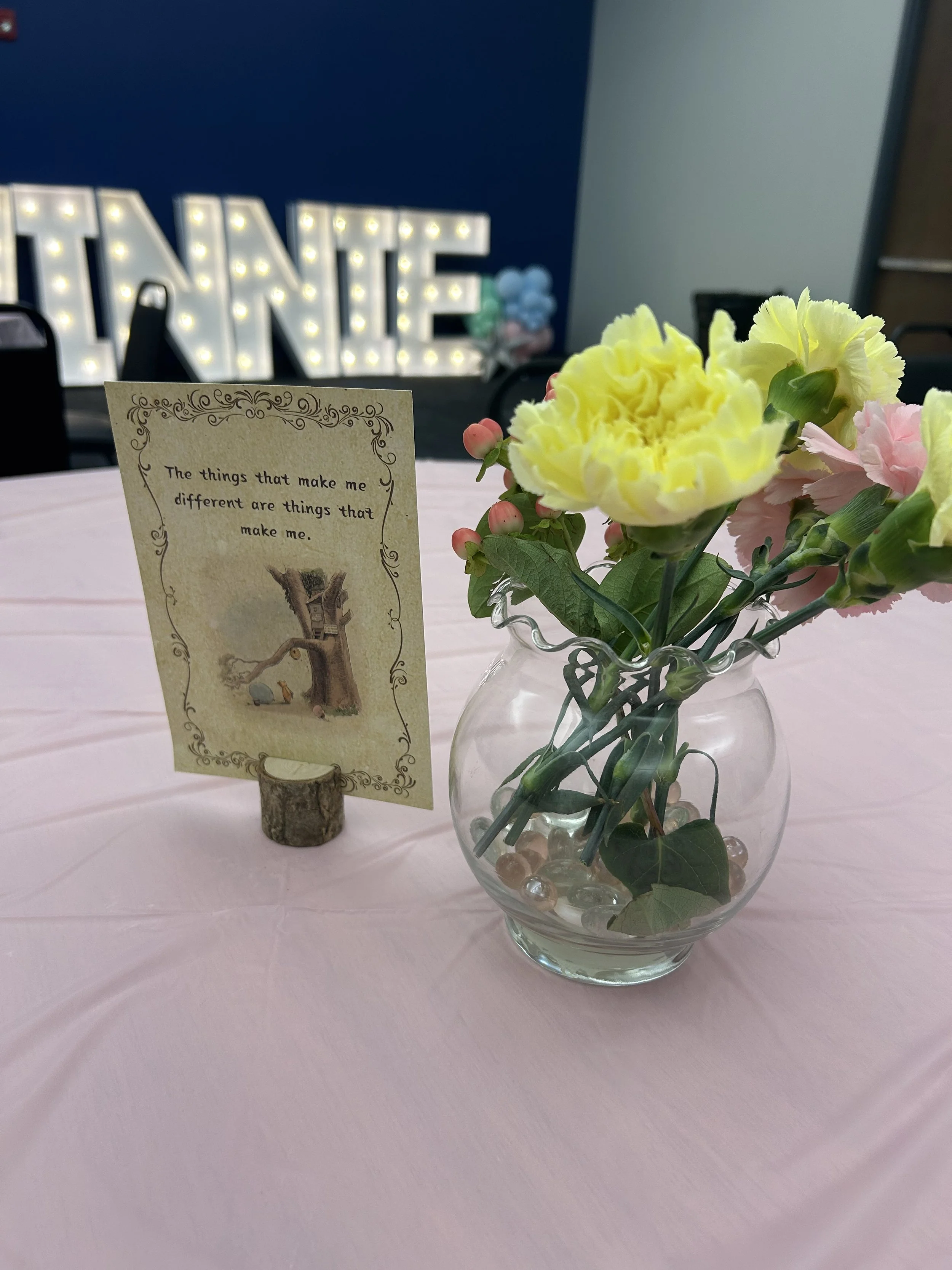 A clear glass vase with yellow and pink flowers on a pink tablecloth, with a decorative card and a illuminated 'winnie' sign in the background.