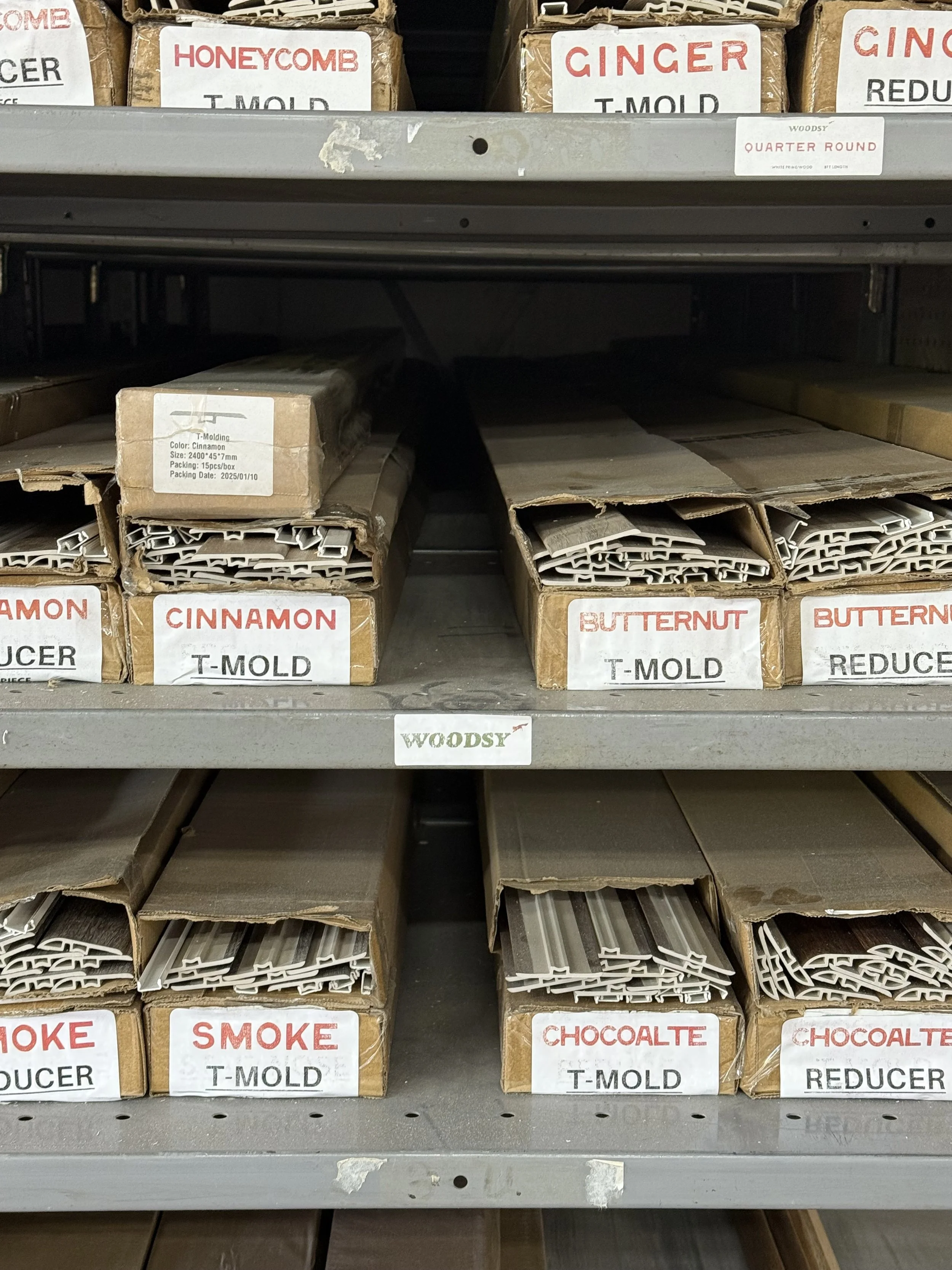 Shelf with wood moldings labeled for different wood types like honeycomb, ginger, cinnamon, butternut, smoke, and chocolate, all in cardboard boxes with white labels.