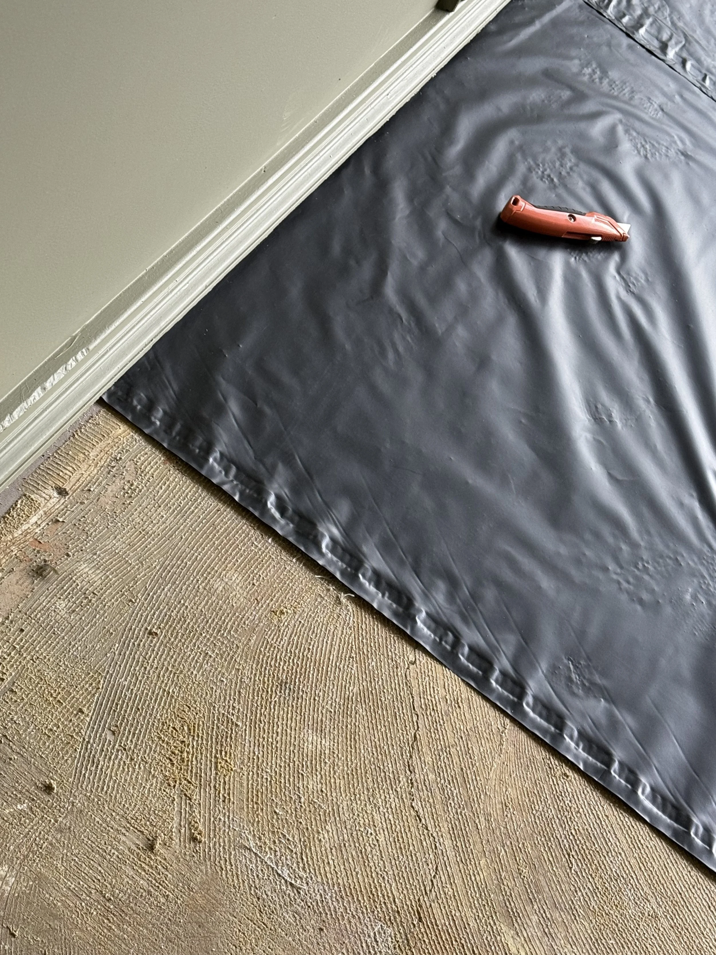A construction adhesive gun resting on a black plastic sheet on a wooden floor, next to a beige wall with white trim.