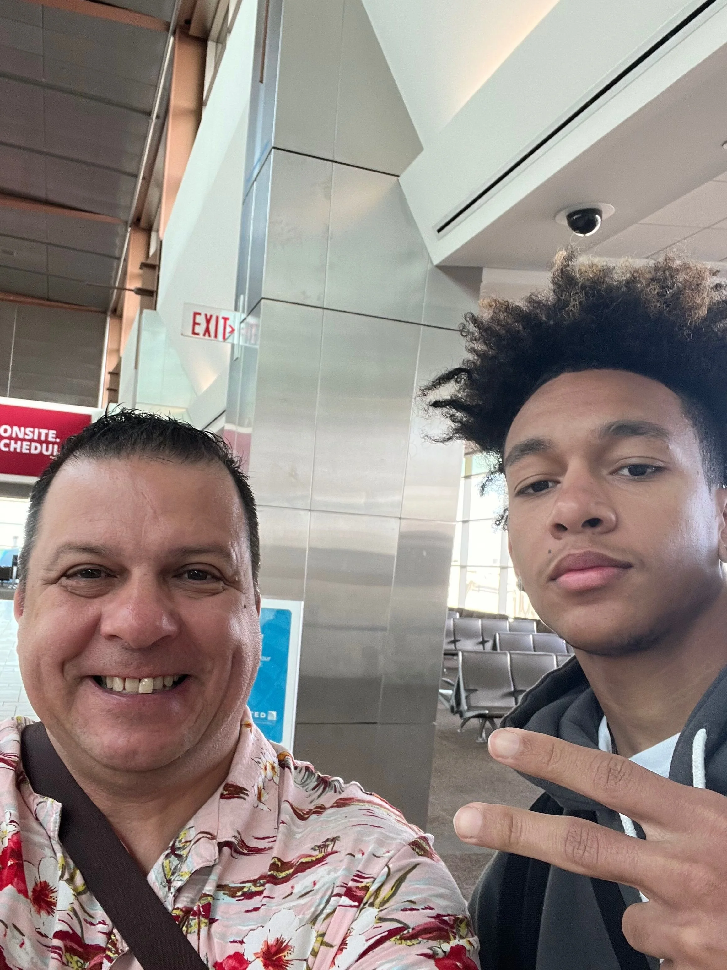 Two men taking a selfie at an indoor public space, with one making a peace sign. The man on the left has short dark hair and is wearing a floral shirt, smiling. The man on the right has curly hair and is wearing a black hoodie, making a peace sign wi
