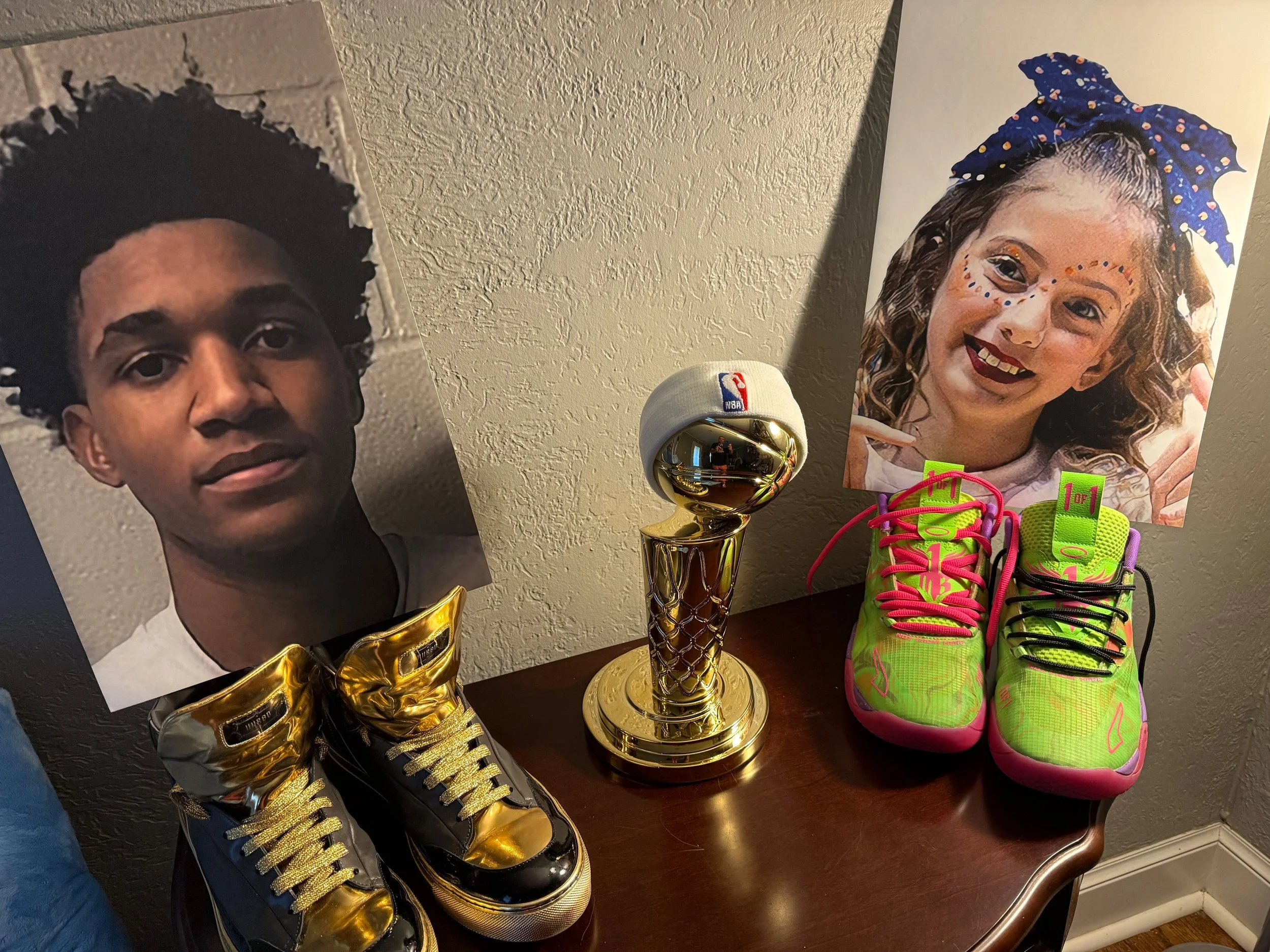 Photographs of a young boy and girl on posters, with a basketball trophy and colorful sneakers on a wooden table.