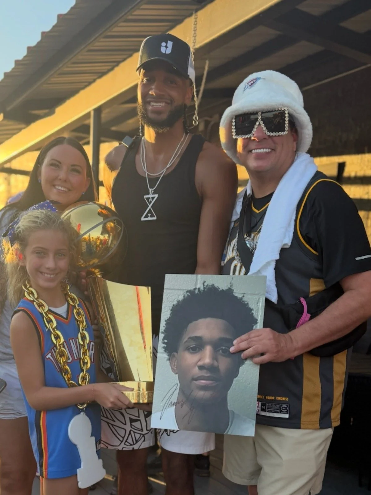 Group of people celebrating, with a man holding a signed photo of a young man with curly hair, a girl wearing a blue basketball jersey with gold chain, a woman, and another man wearing sunglasses, a white fur hat, and a towel around his neck. The per