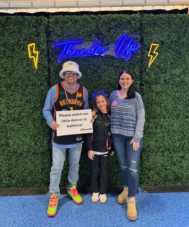 Three people standing in front of a green hedge wall with neon sign 'Thunder Up' and yellow lightning bolts. The man on the left is wearing a basketball jersey, hat, and colorful sneakers, holding a sign that says 'Please watch our little dancer at h