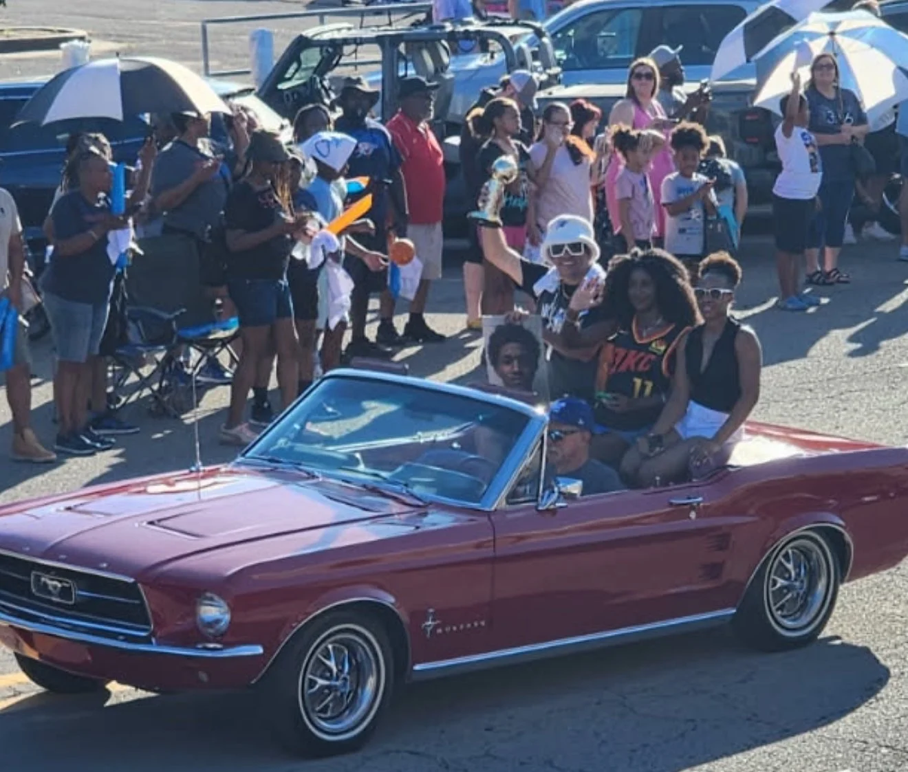 A red vintage Ford Mustang convertible driving through a parade with a crowd of spectators on the side of the street, some holding umbrellas for shade.