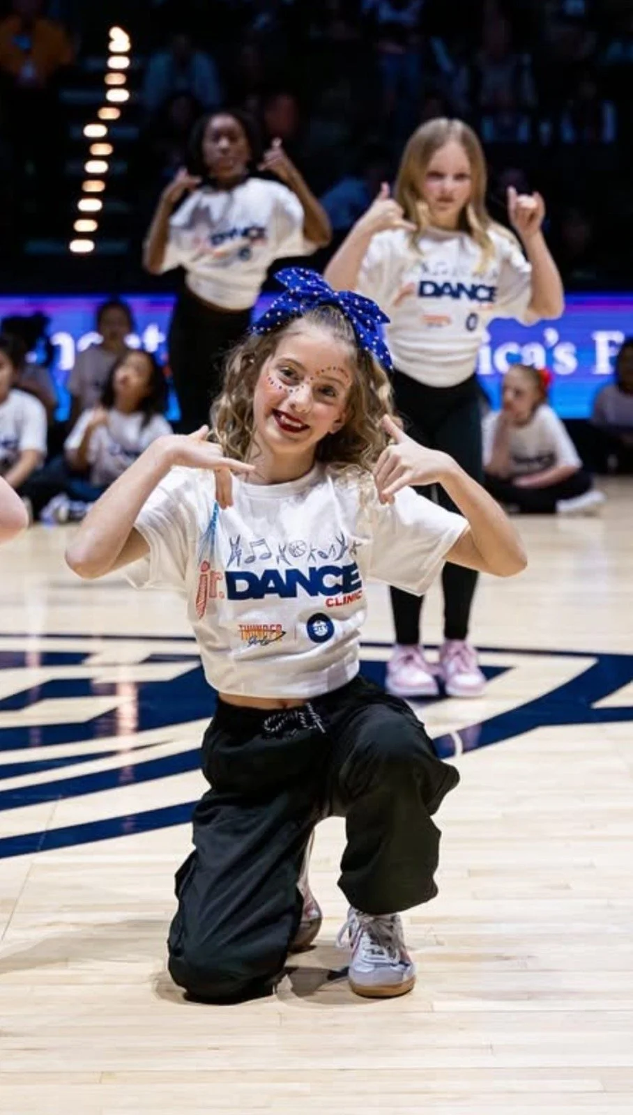 A young girl with curly hair, face paint, and a blue bow on her head is posing on a dance floor with her fingers pointing to her head. She is wearing a white T-shirt with the word 'DANCE' on it and black pants. Behind her, other children in matching