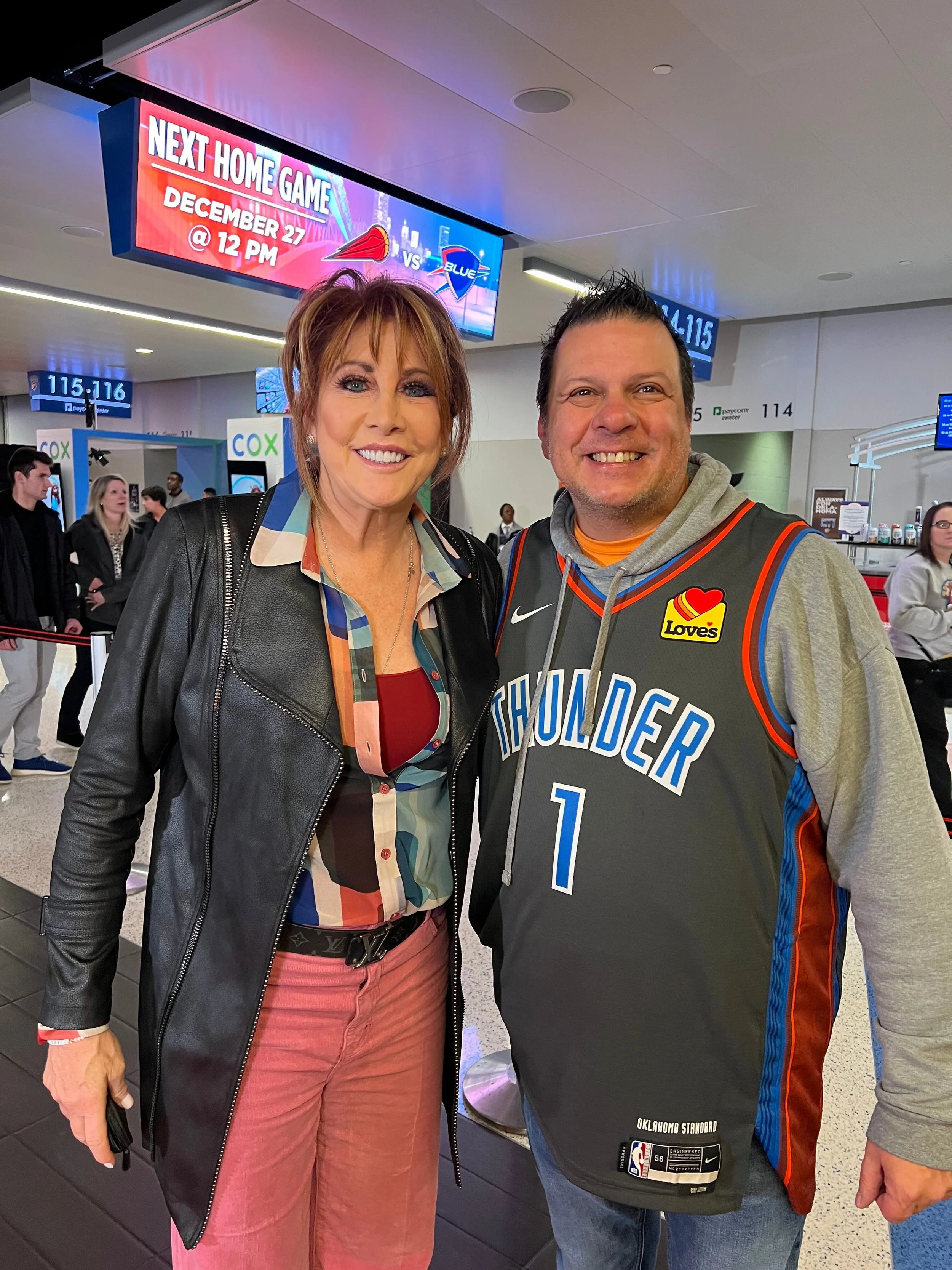 A woman and a man smiling at the airport, standing in front of a digital sign advertising a basketball game between the Thunder and the Blue, scheduled for December 27 at 12 PM, with a group of travelers in the background.