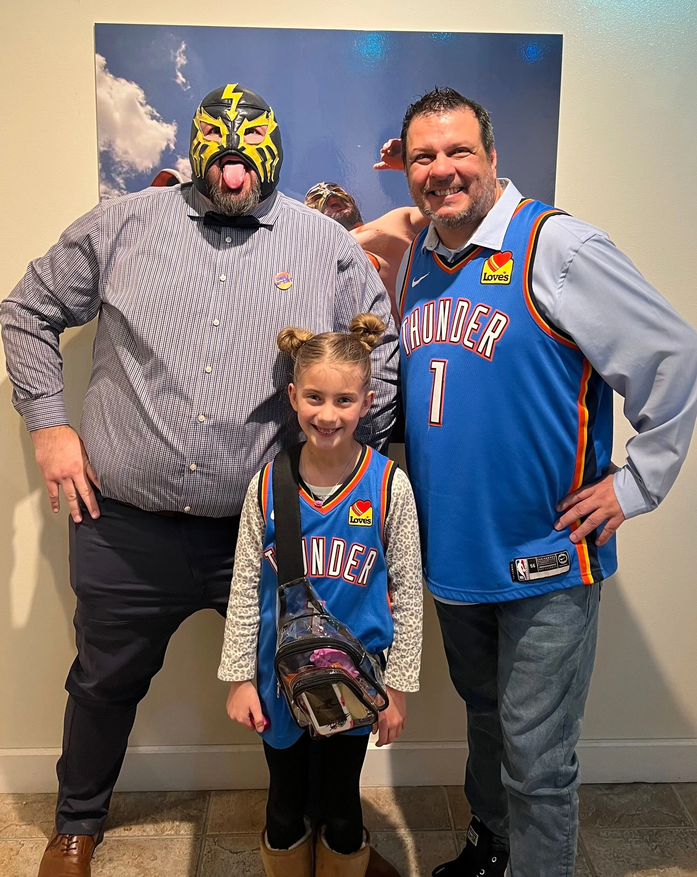 A young girl wearing a Thunder basketball jersey and carrying a backpack stands with two men and a person wearing a luchador mask, posing together indoors against a painted sky background.