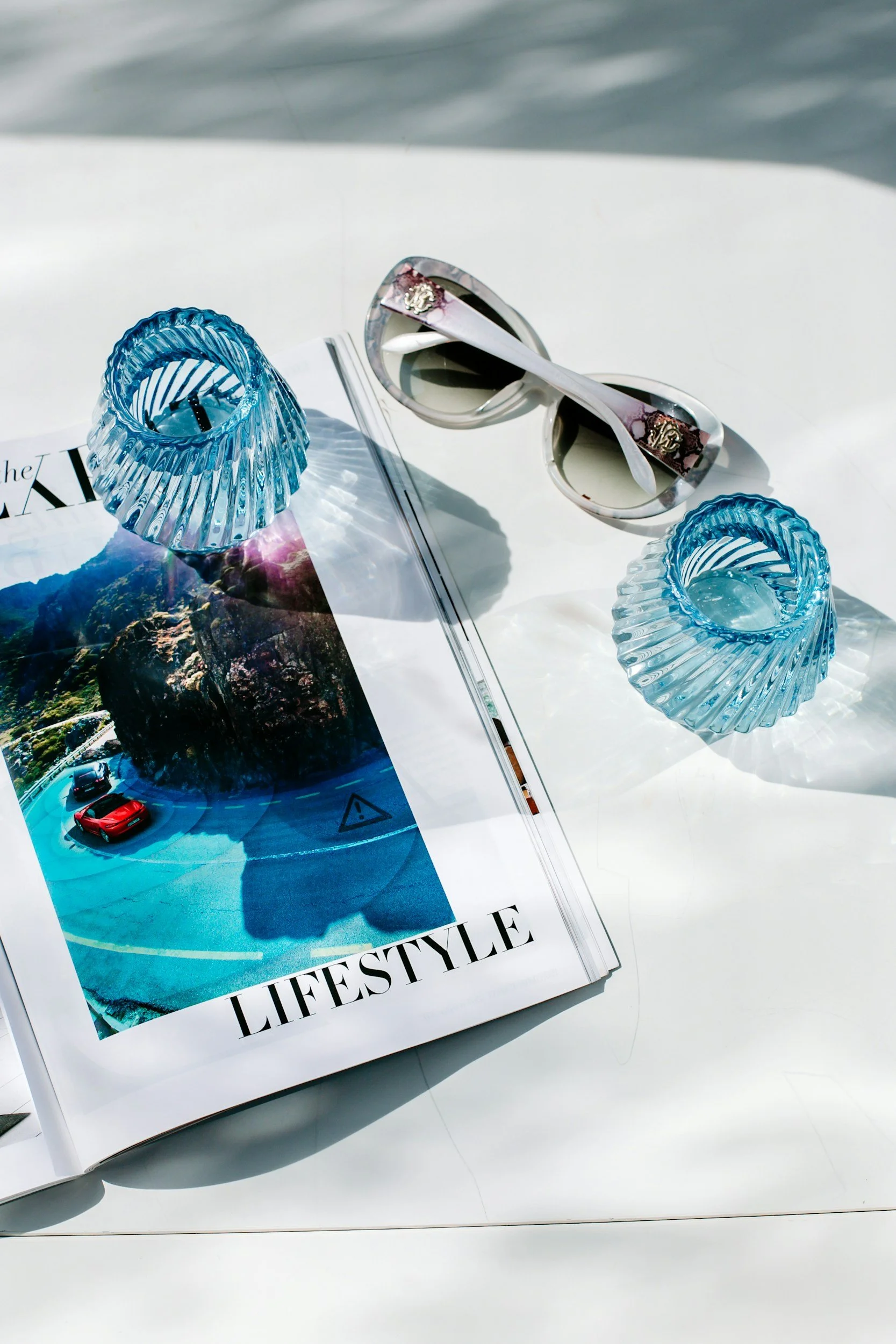 A white table with a magazine, two blue glass candle holders, a pair of white sunglasses with a floral design, on a sunny surface.