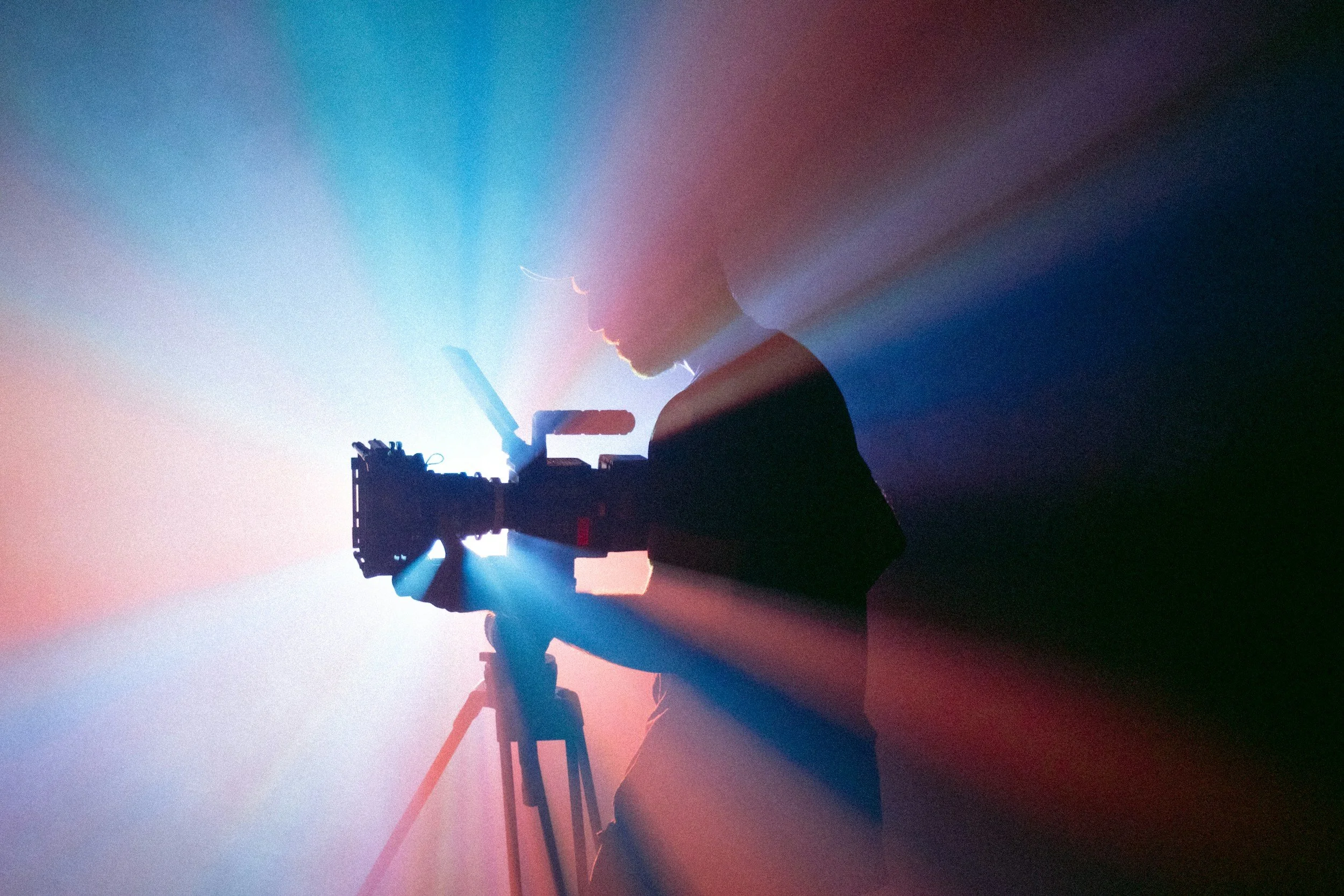 Silhouette of a person operating a camera or filming equipment, with colorful light rays streaming outward.