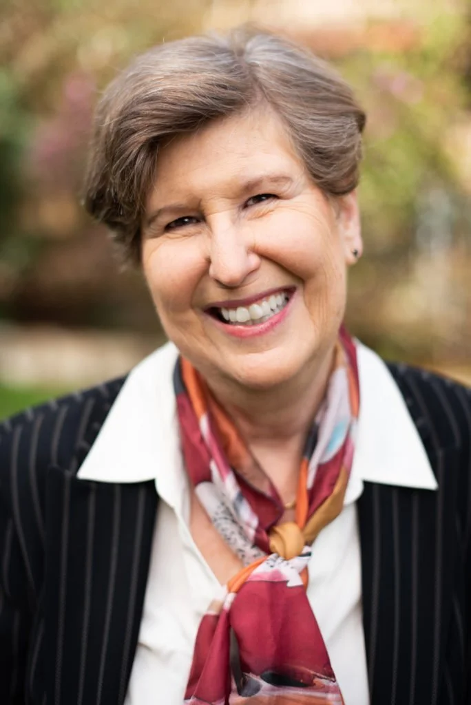 Smiling older woman with short gray hair, wearing a white shirt, black pinstripe blazer, and a colorful scarf, outdoors with blurred autumn trees in the background.