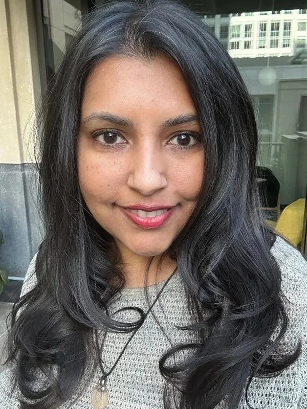 Close-up of a woman with long, dark, wavy hair smiling at the camera, wearing a light gray sweater and a necklace, with a cityscape background