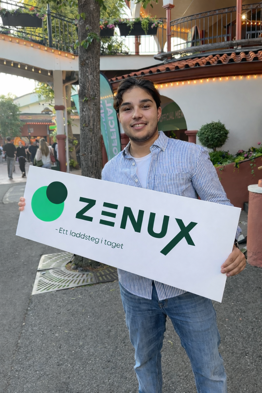 En lycklig ung man håller ett reklamskylt för Zenux, ett laddsteg i taget, utomhus på ett torg eller gatustråk, omgiven av andra människor och byggnader med belysning.