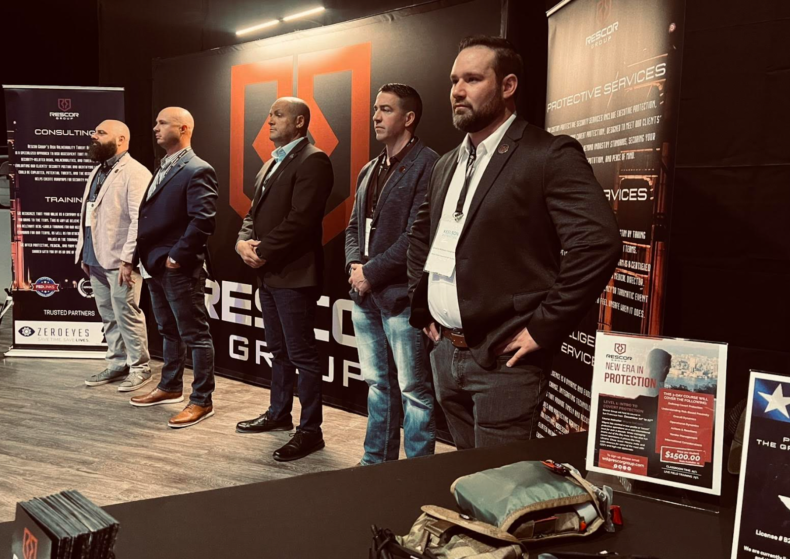 Six men standing in a row at a conference or event, with banners and informational displays in the background. They are dressed in business or business-casual attire.