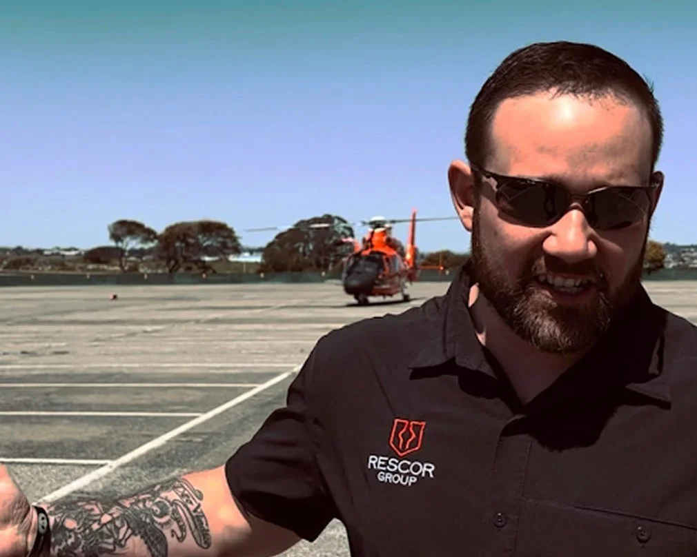 Man wearing black Rescor Group shirt and sunglasses standing in front of a helicopter on a tarmac with trees and blue sky in the background.