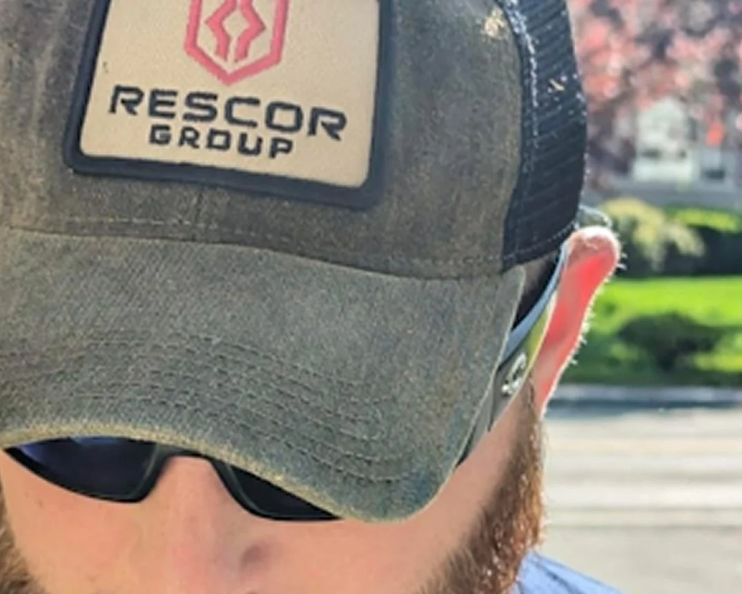 Close-up of a person wearing a gray baseball cap with a 'Rescor Group' logo, dark sunglasses, and earbuds, outdoors with a blurred street and trees in the background.