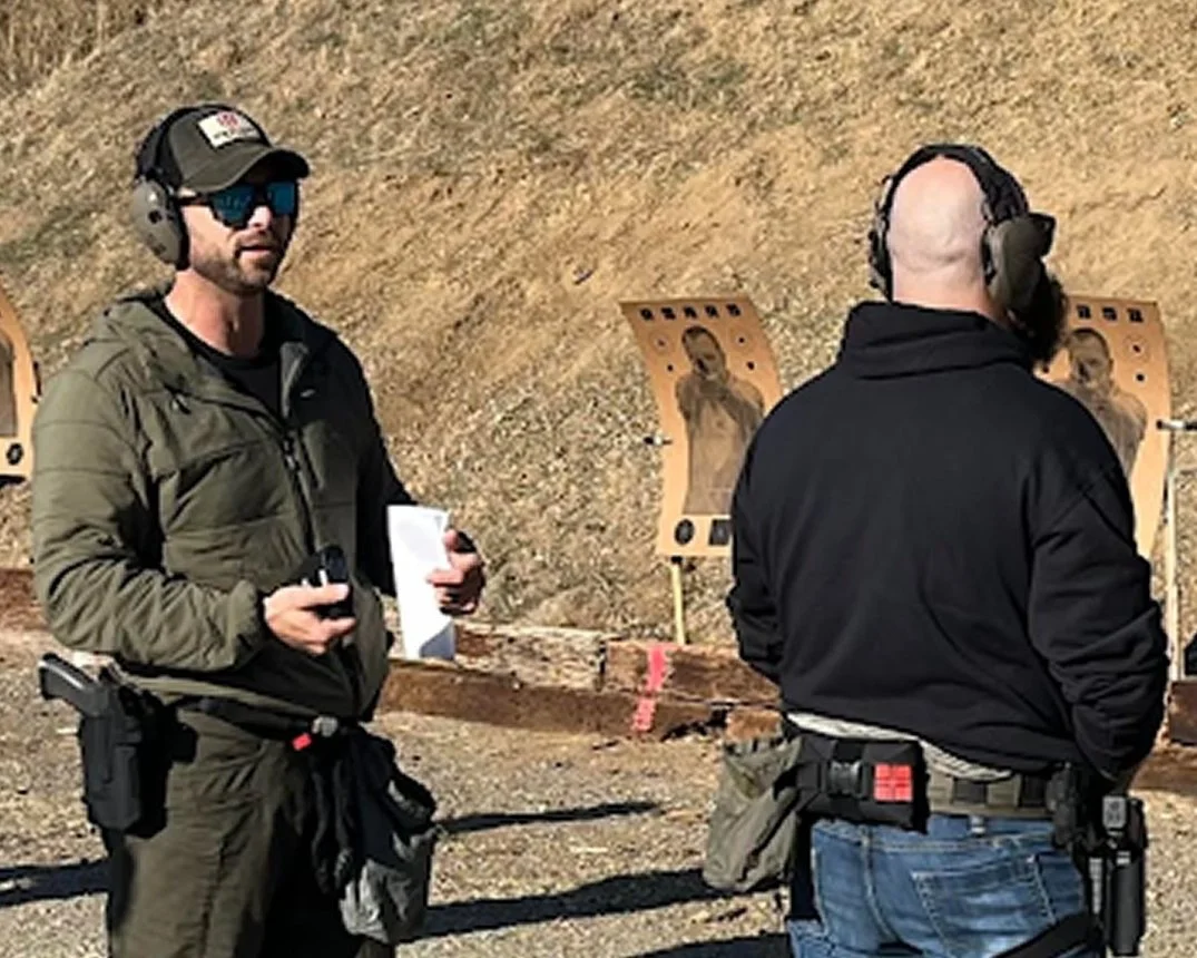 Two men wearing protective hearing and eye gear at outdoor shooting range, with targets depicting a person.