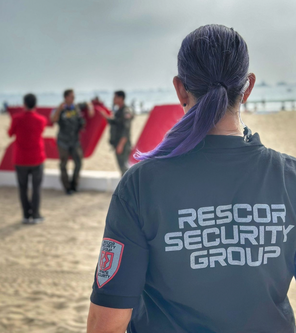 A security guard with purple hair, wearing a black shirt with 'Rescor Security Group' on the back, is watching a group of people setting up red letters on a sandy beach. In the background, the ocean and cloudy sky are visible.