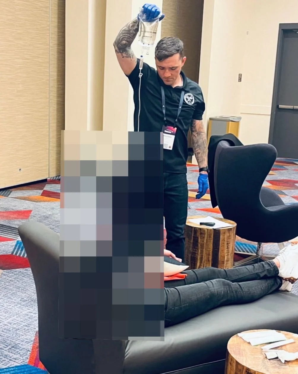 A person lying on a couch receiving medical assistance from a man with gloves holding an IV bag.