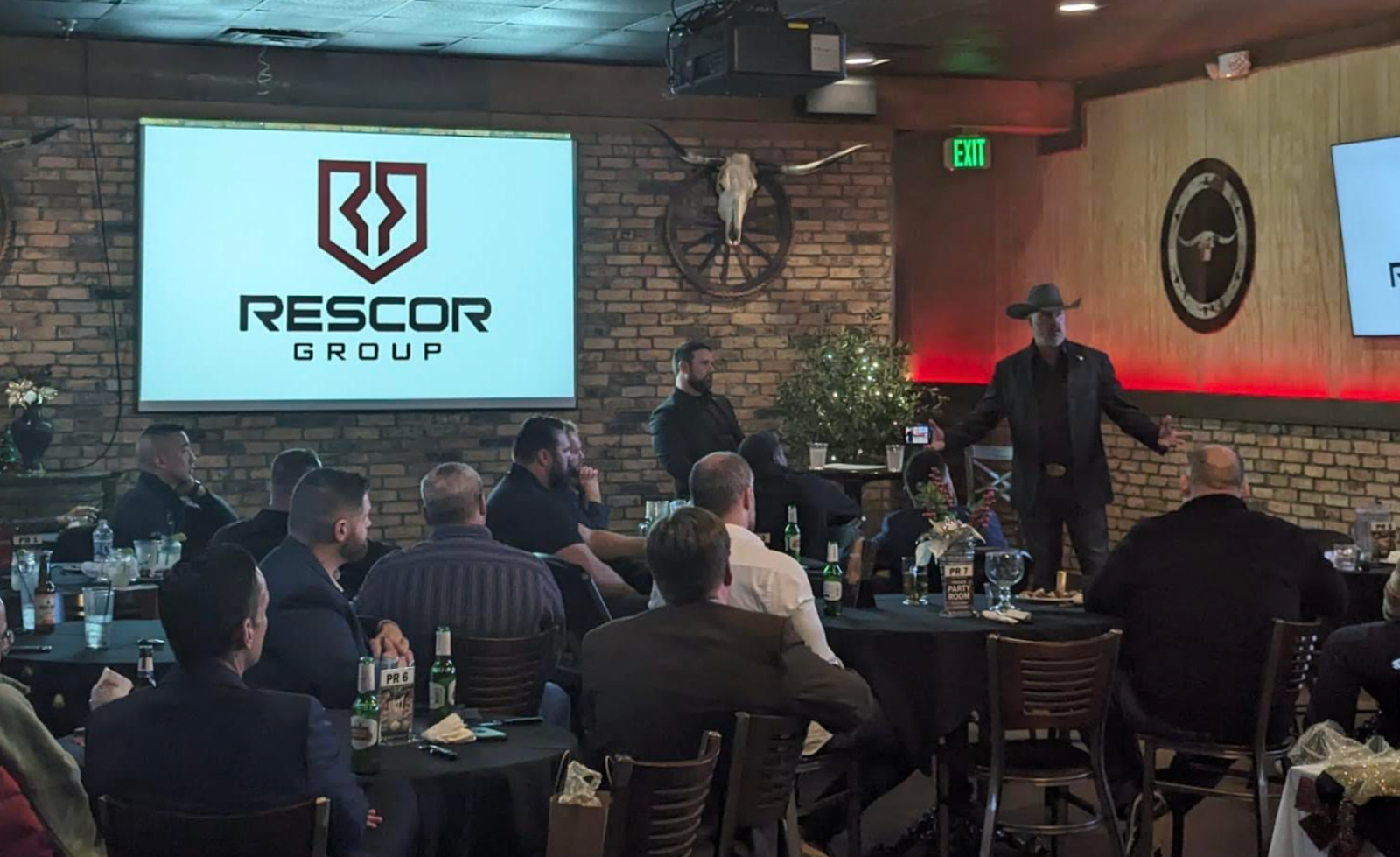 A man in a cowboy hat giving a presentation in a restaurant or event space with a brick wall, a large screen displaying 'Rescor Group' logo, and seated audience members.