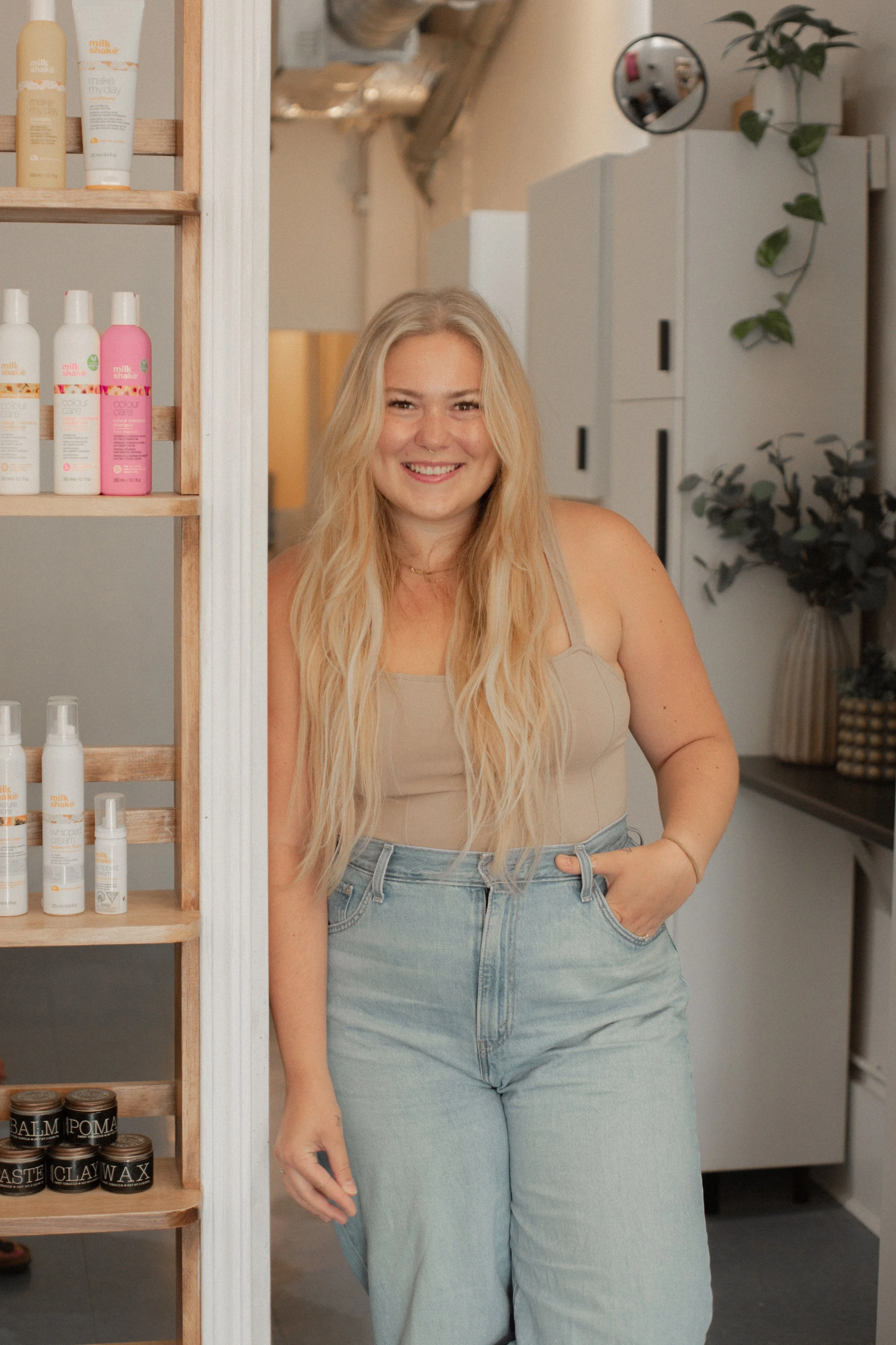 Elyssa with long blonde hair smiling and standing next to a wooden shelf filled with skincare and haircare products in a modern, cozy interior.