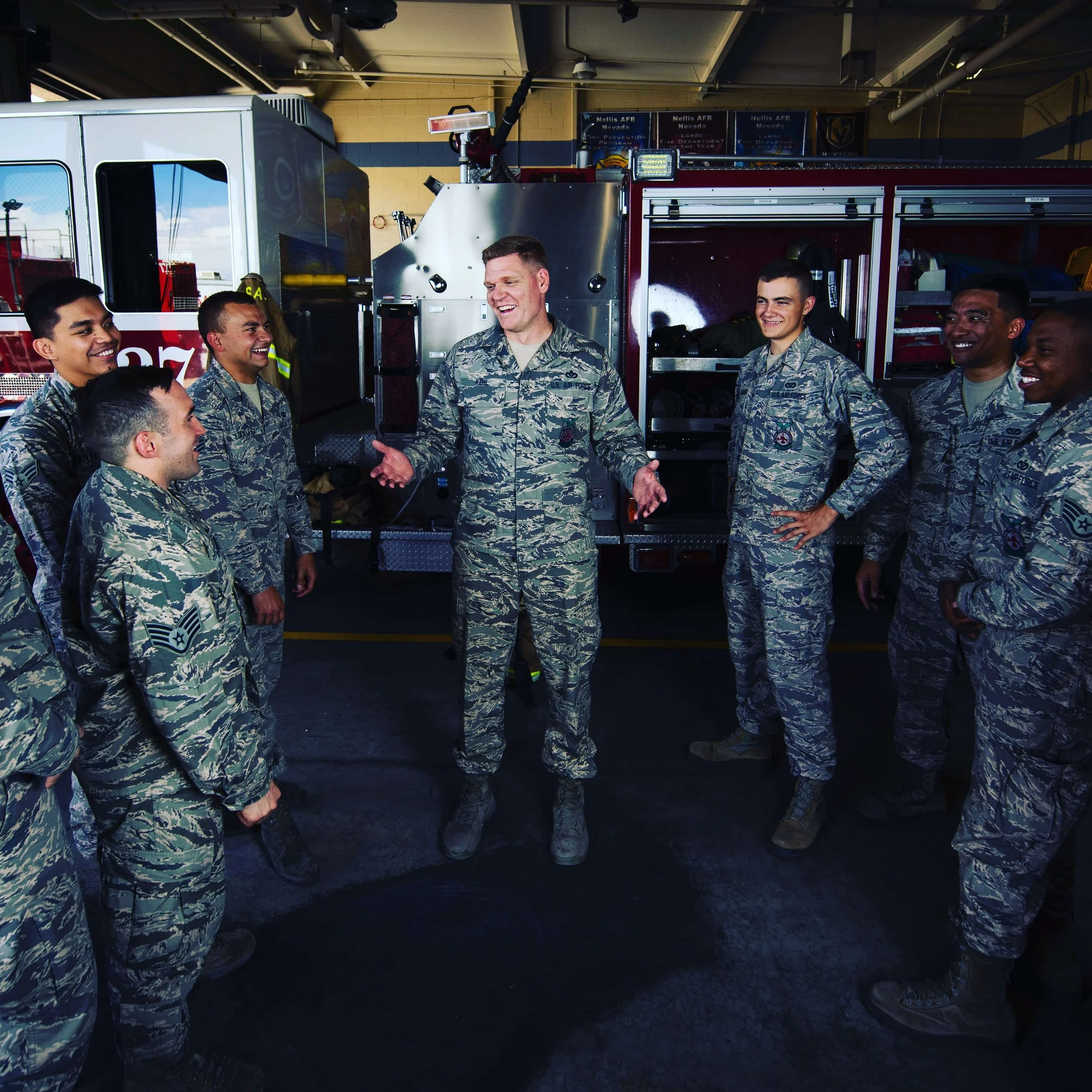 A group of military personnel gathered inside a fire station, listening to a smiling officer who appears to be giving instructions or a briefing.