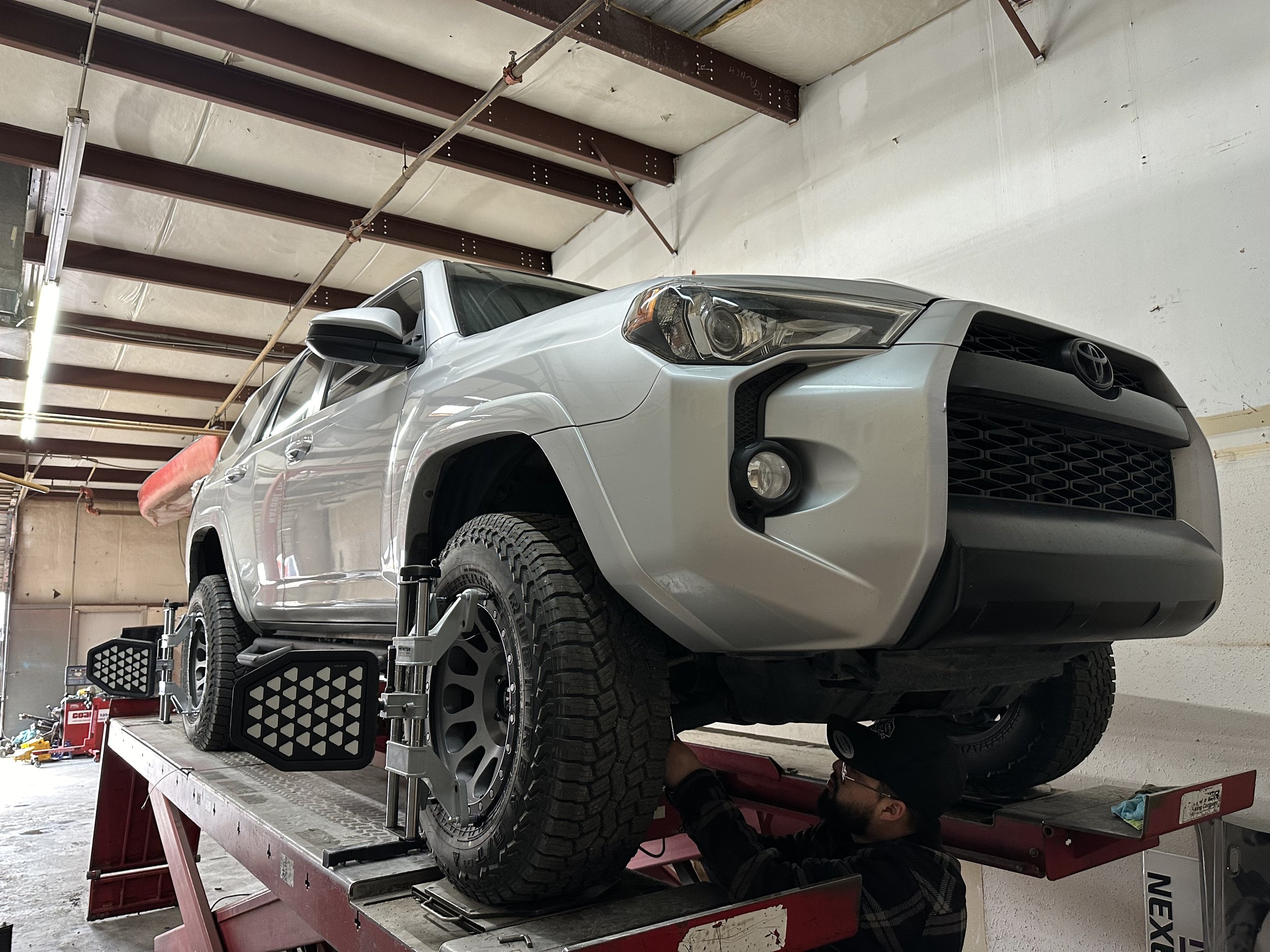 A silver pickup truck is elevated on a hydraulic lift in an auto repair shop, with a mechanic working underneath it.