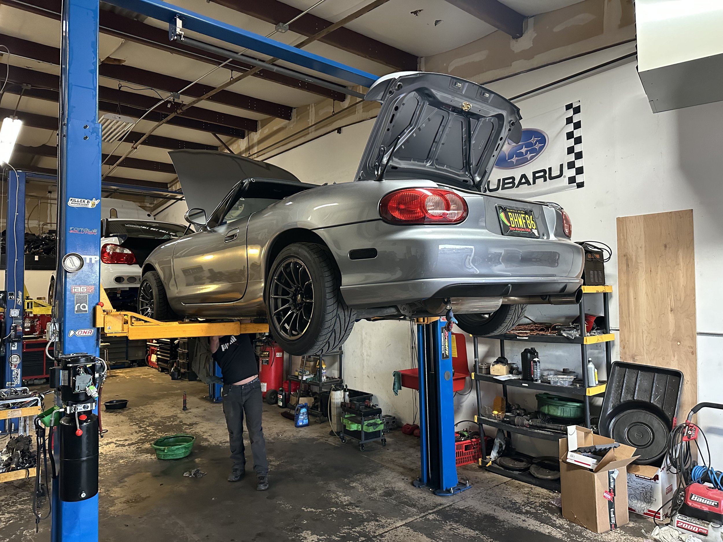 Inside an automotive garage, a silver Subaru convertible with its hood and trunk open is raised on a hydraulic lift. A mechanic, dressed in black, is working underneath the car. The garage has tools, parts, and storage shelves, with a Subaru banner on the wall.