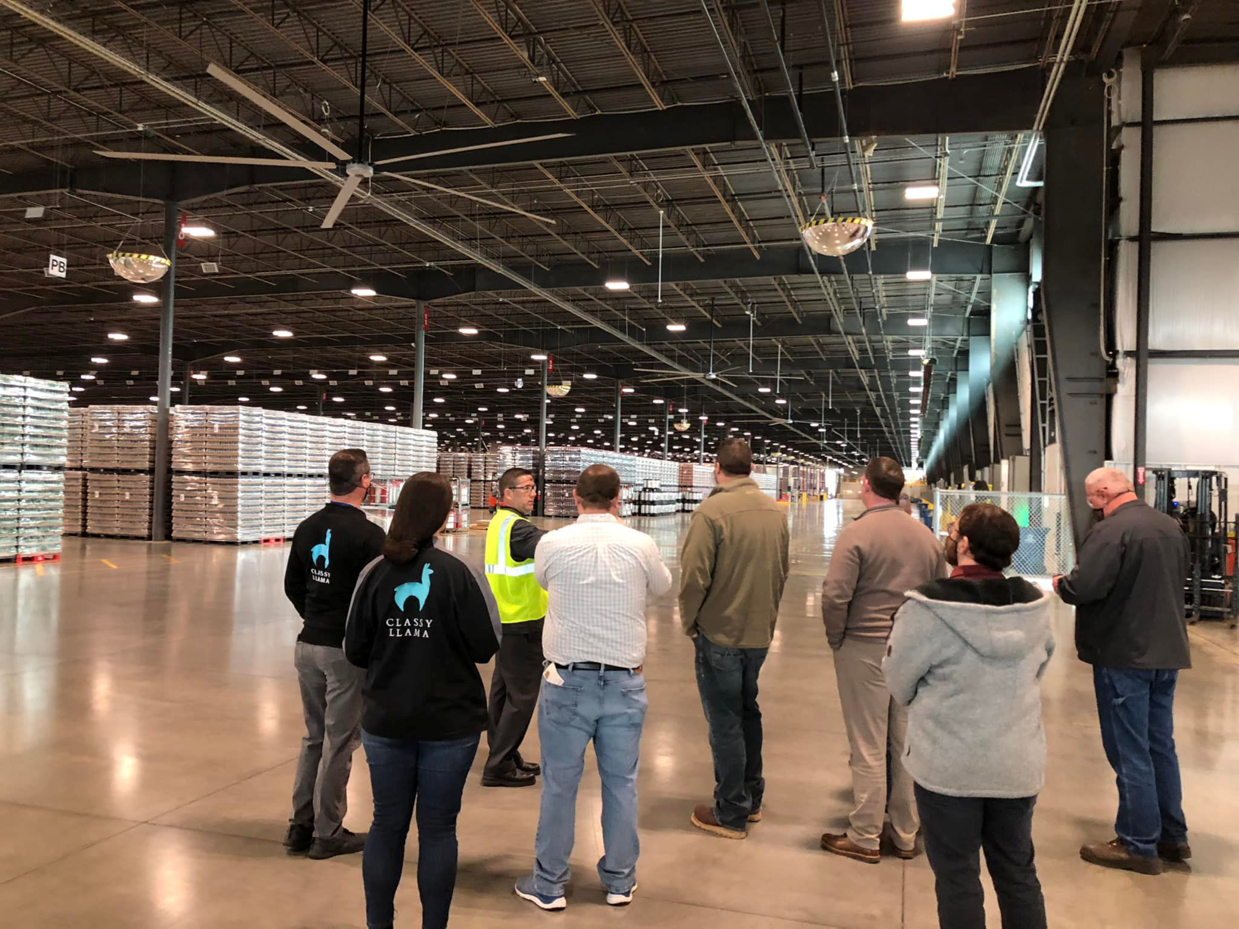 Group of people touring a large warehouse, some wearing branded jackets with a llama logo, looking at stacked pallets of goods.