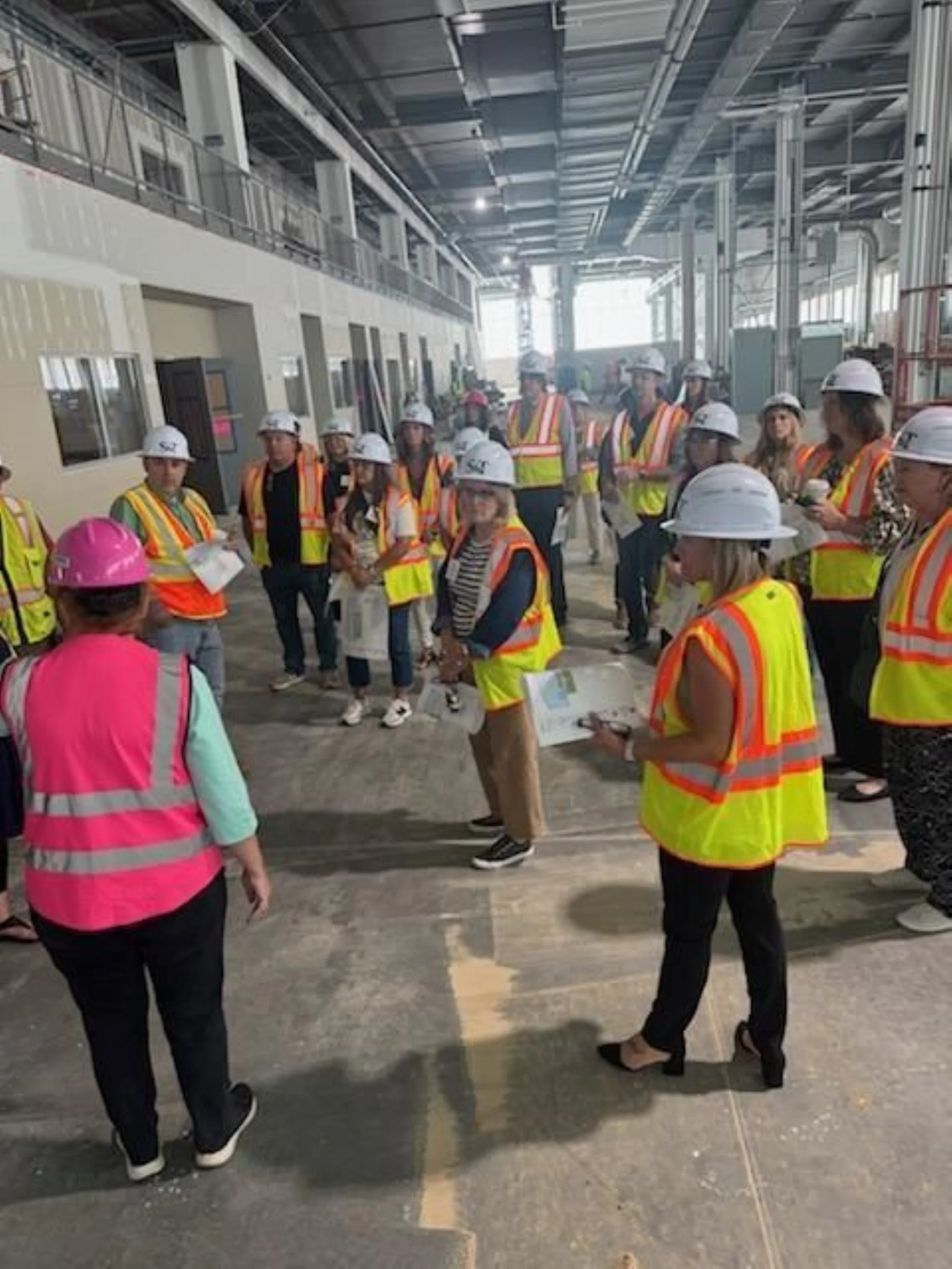 A group of construction workers and supervisors wearing safety vests and helmets gather inside a large building under construction, listening to a person speaking.