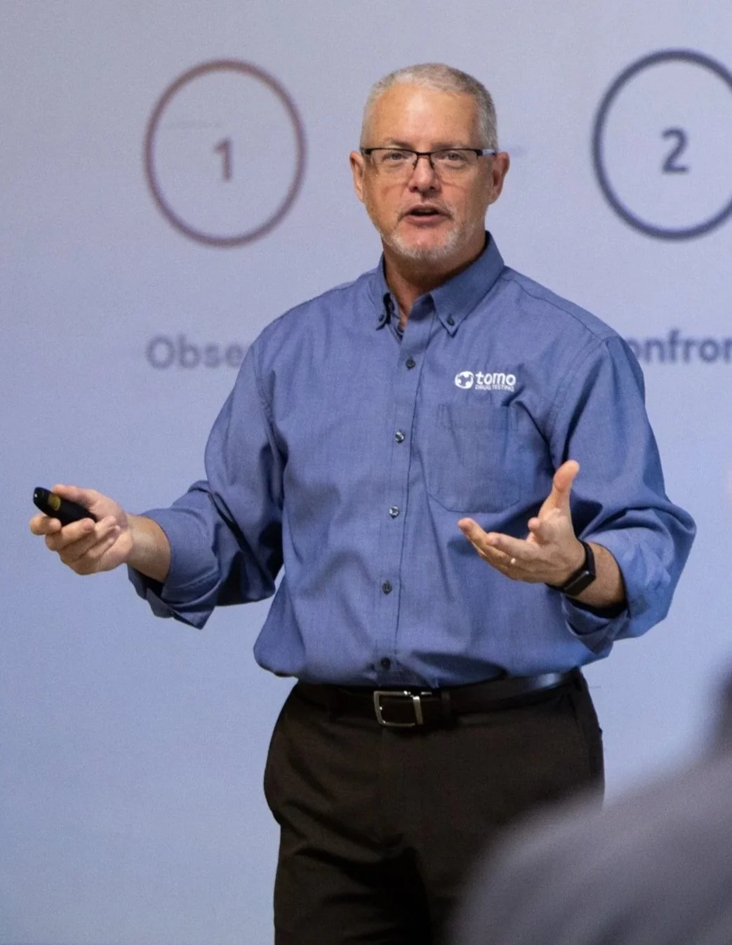 A man with glasses and gray hair, wearing a blue shirt with a logo, is giving a presentation with a remote in his hand and a background of a projected slide showing numbered circles.