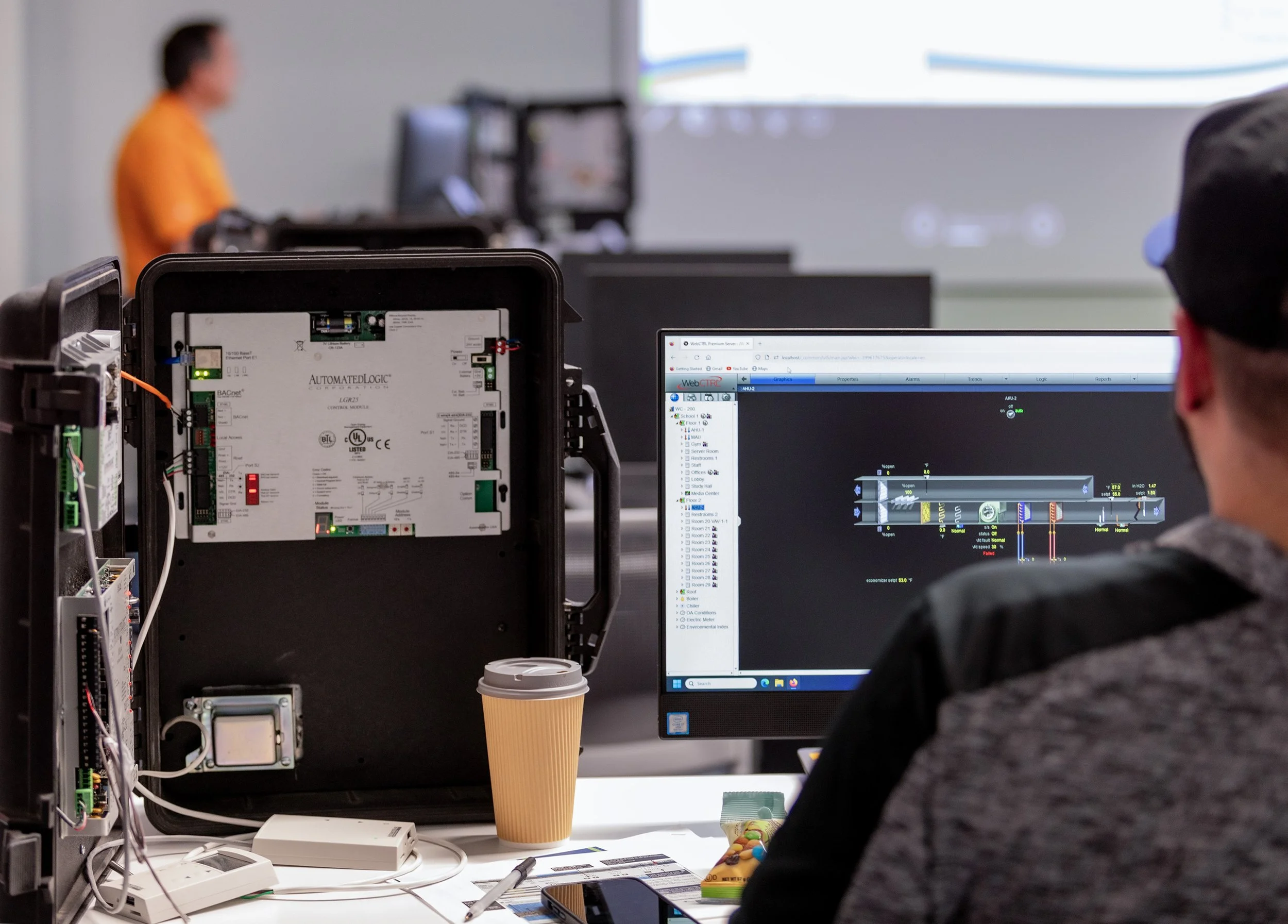 A person sitting at a desk with a computer monitor displaying a diagram or schematic, with open electronic components and a coffee cup on the desk. In the background, another person is working at a separate computer.