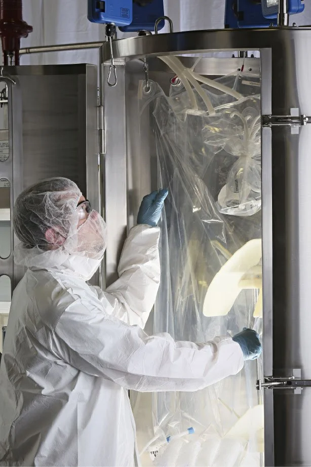 A scientist in full protective gear, including a white gown, gloves, and a face mask, working inside a biosafety cabinet with clear plastic cover, handling laboratory equipment.