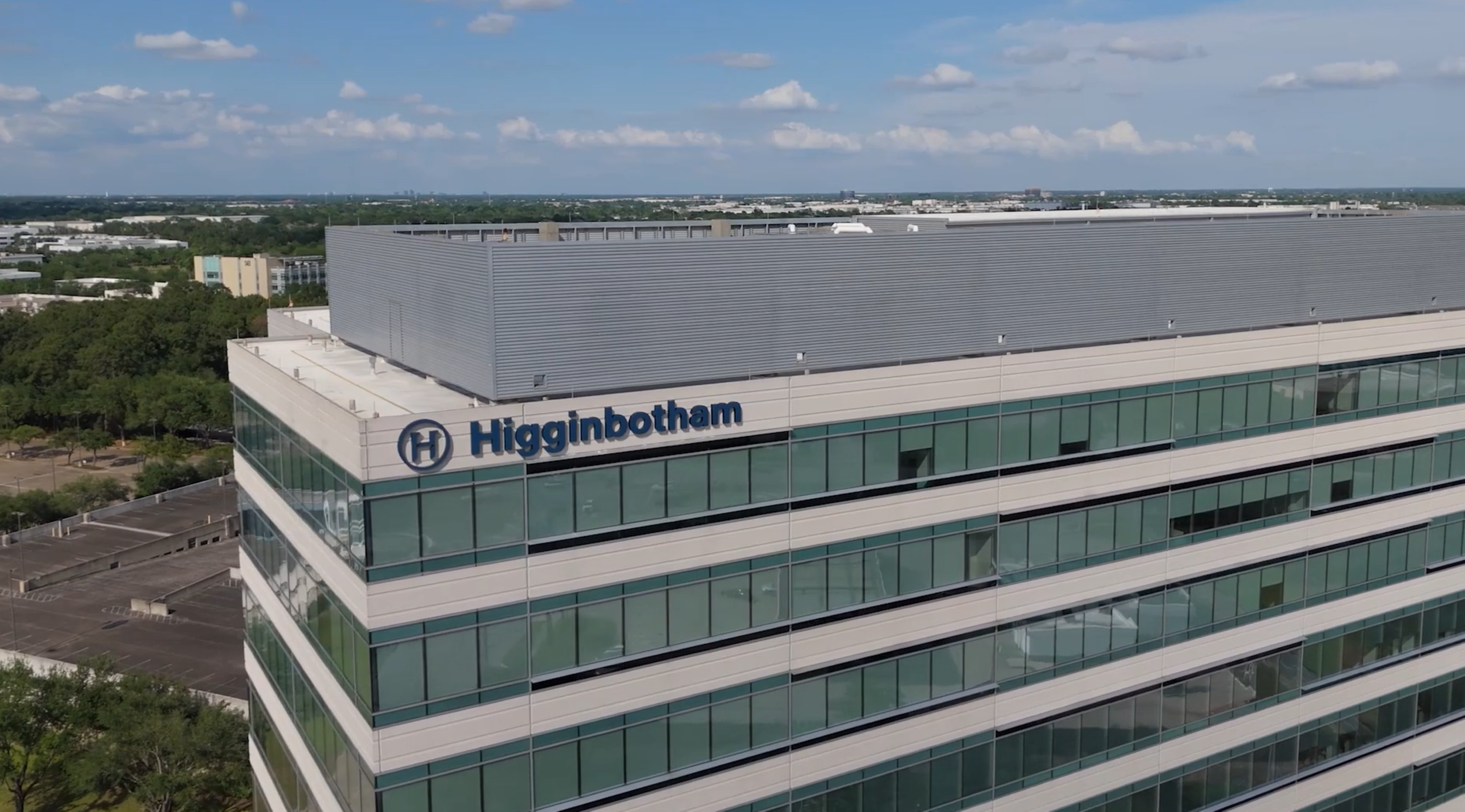 A tall modern office building with the logo and name 'Higinbotham' on the top corner, with several floors of glass windows, surrounded by trees and parking lots under a partly cloudy sky.