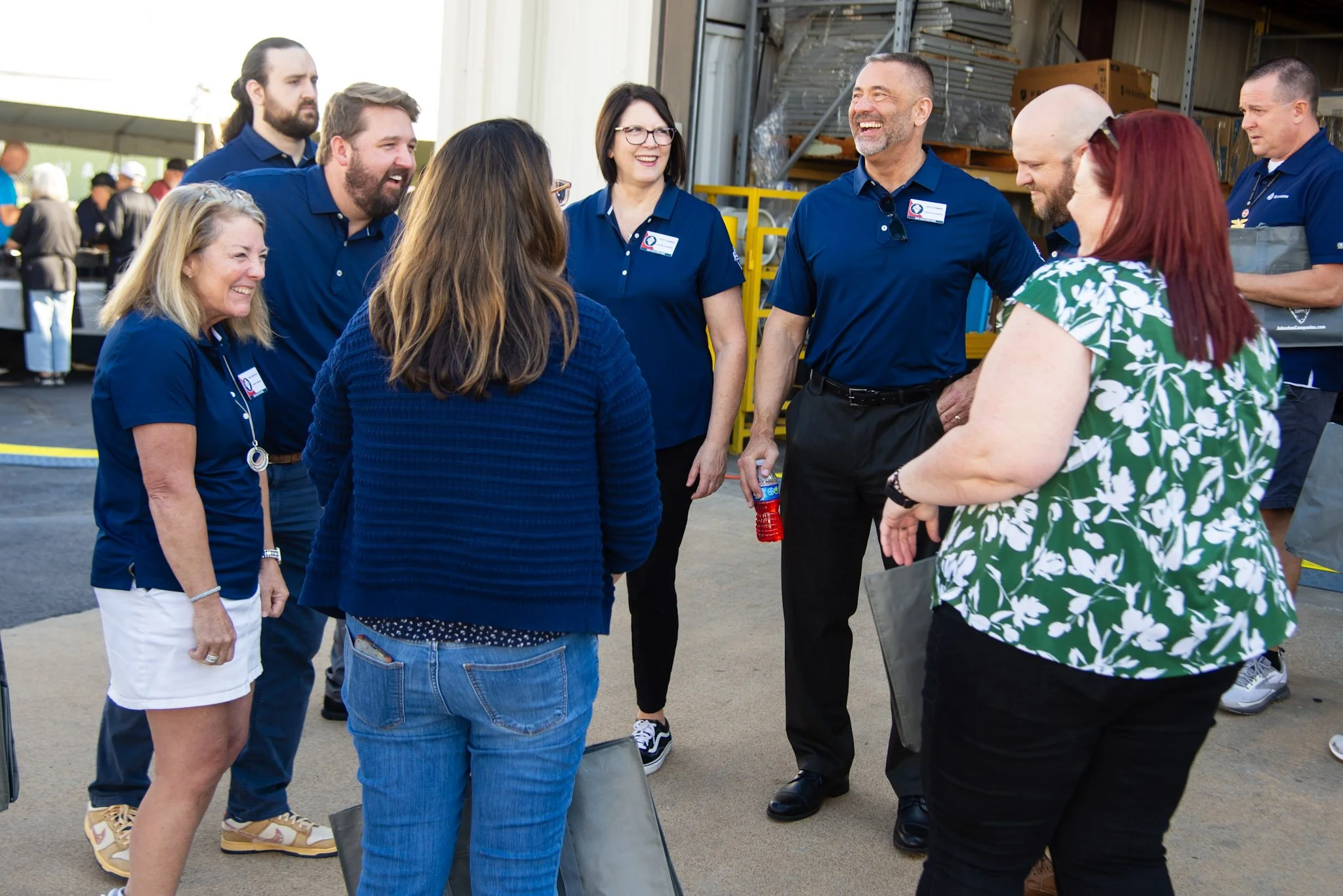 Group of people standing and talking outside a warehouse, some wearing matching navy shirts with name tags, appearing to enjoy a conversation.