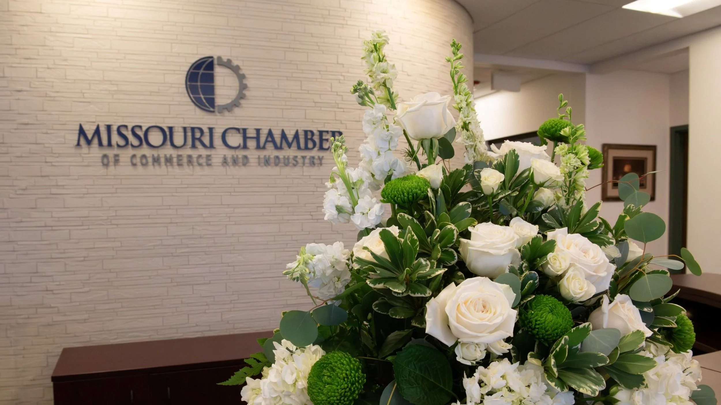 White floral arrangement with roses, hydrangeas, and green accents in front of a Missouri Chamber of Commerce and Industry sign.