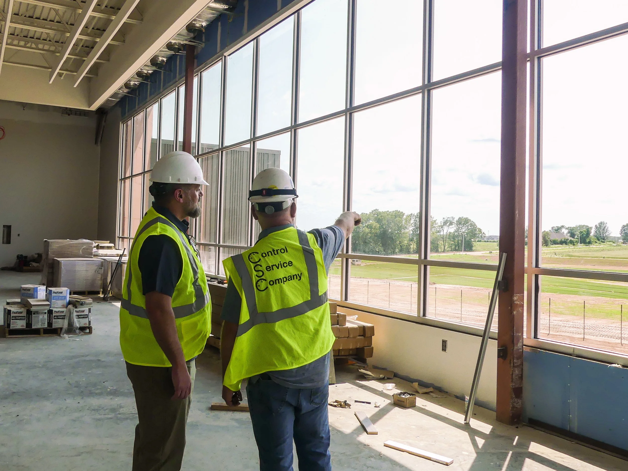 Two construction workers in yellow safety vests and white helmets standing indoors near large window, discussing construction progress while pointing outside.