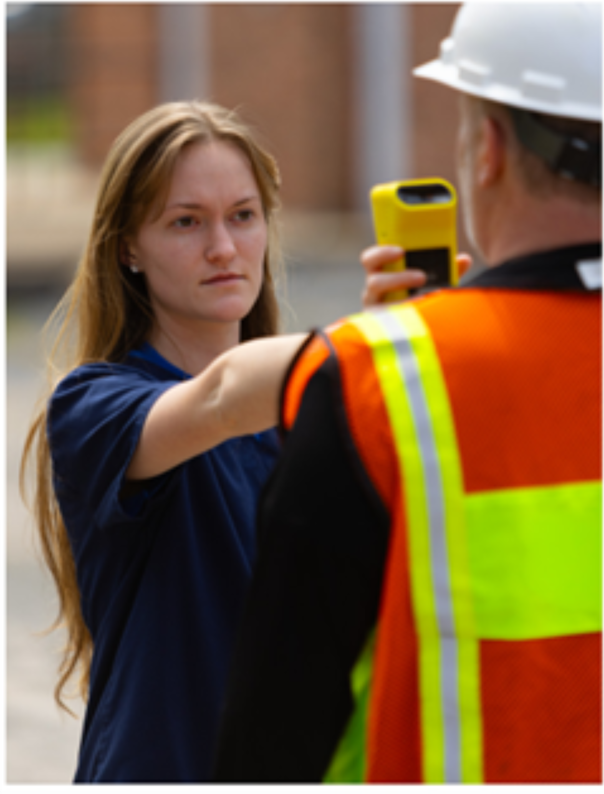 A young woman with long hair and a serious expression is taking a photo or recording a video of a man wearing a safety vest and a hard hat with a yellow phone.