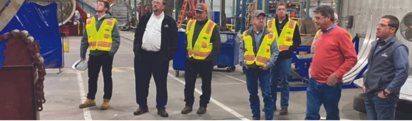 Group of men in safety vests and casual clothing standing inside industrial or warehouse setting, some with hands in pockets, listening to a speaker or presentation.