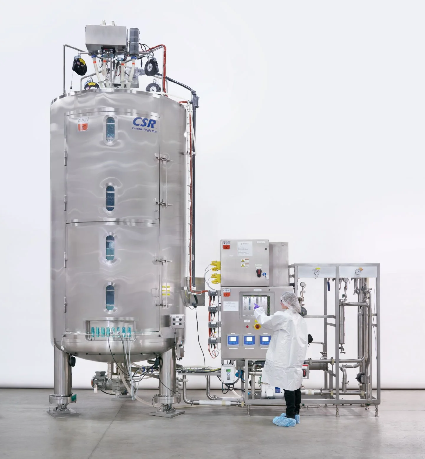 A scientist operating machinery in a clean lab environment. The large stainless steel vessel has the logo 'CSR' and labels, with various pipes and gauges connected. The scientist wears protective clothing, including a lab coat, hairnet, gloves, and shoe covers.