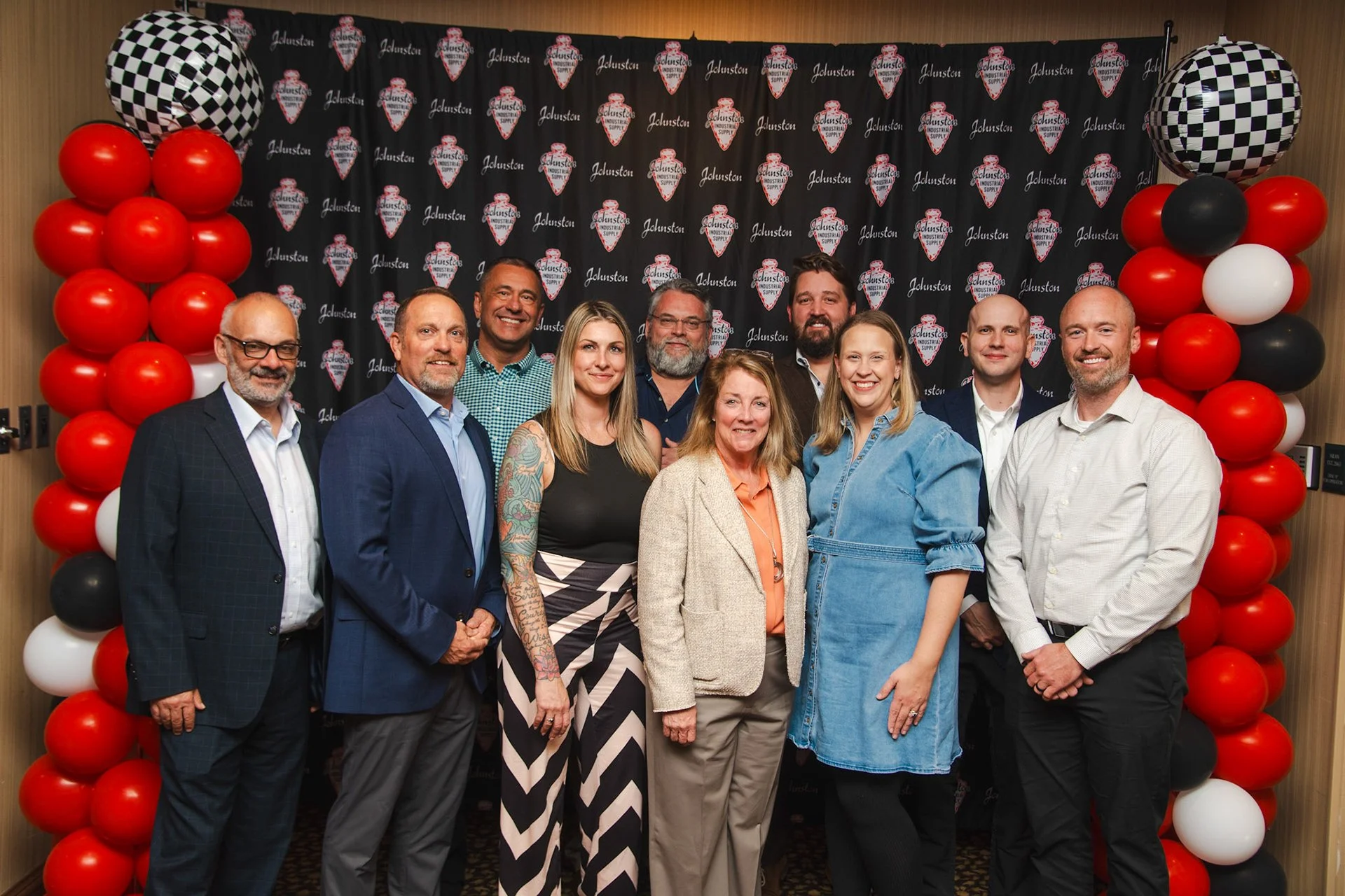 A group of eleven people posing in front of a backdrop with logos, surrounded by red, white, black, and checkered balloons at an event or celebration.