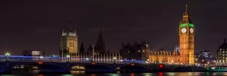 Nighttime view of the Big Ben clock tower and the Palace of Westminster in London, England, illuminated and reflecting on the river.