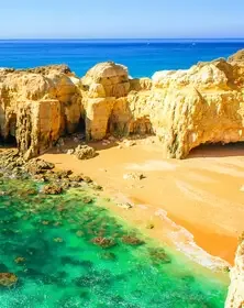 A beach with golden sand, large yellow rock formations, and clear turquoise water, under a blue sky.