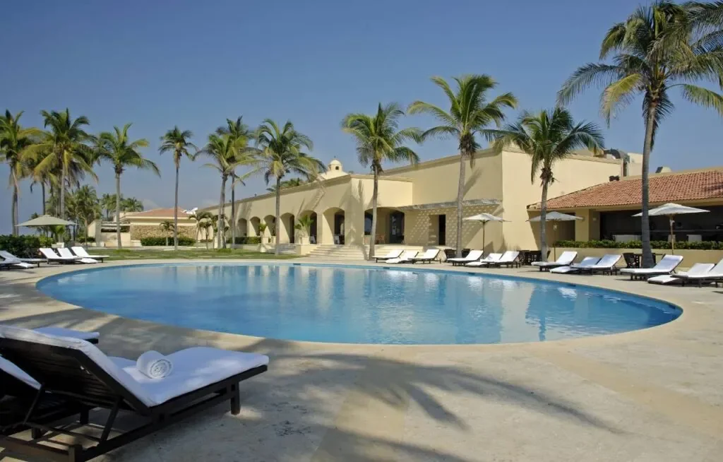 A tropical resort pool area with palm trees, lounge chairs, and a building in the background under a clear blue sky.