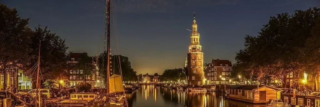 Night view of a city canal with boats, illuminated buildings, and a tall historic tower in the background.