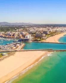 Aerial view of a beach with a pier extending into the ocean, cityscape in the background, blue sky, and clear turquoise waters.