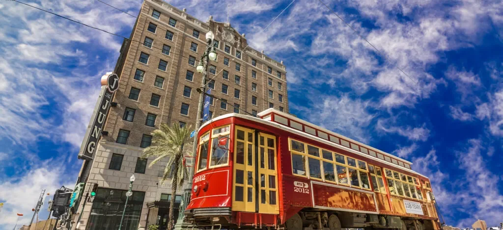 A red and yellow vintage trolley bus on a city street, with a high-rise building and palm trees in the background, under a partly cloudy sky.