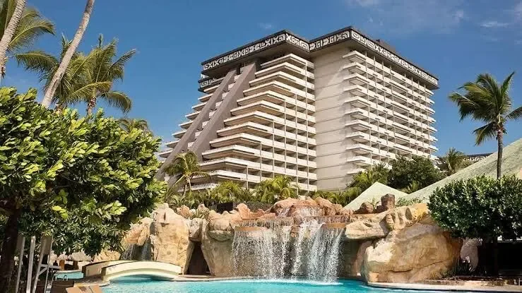 A tall hotel building with multiple balconies surrounded by palm trees and greenery. In front, there is a swimming pool with rocks and a waterfall feature, all set in a tropical resort setting.