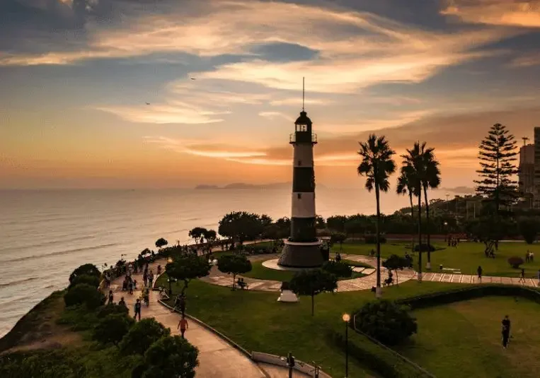 Lighthouse on a grassy park by the ocean during sunset, with palm trees and people walking along the paths.