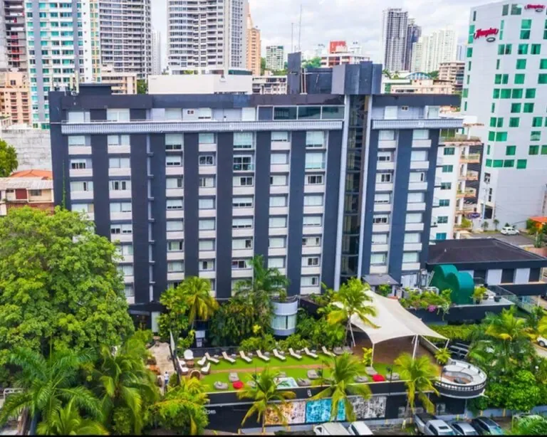 Tall gray hotel building with multiple windows, surrounded by palm trees and outdoor lounge area with chairs and umbrellas, in an urban cityscape.